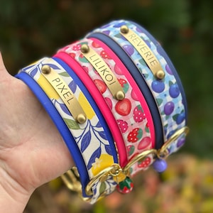 May include: Three pet collars featuring gold-tone metal nameplates engraved with the names PIXEL, LILIKOI, and REVERIE. The collars display patterns of yellow lemons, red strawberries, and blue blueberries on gingham backgrounds with gold hardware.