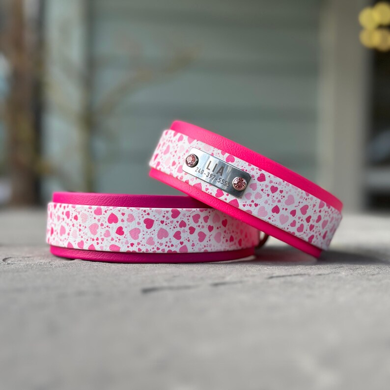 May include: Two bright pink collars featuring a white band with a repeating pattern of small pink and red hearts. The top collar includes a rectangular silver metal identification tag engraved with the name LIA and the phone number 240-397-5555.
