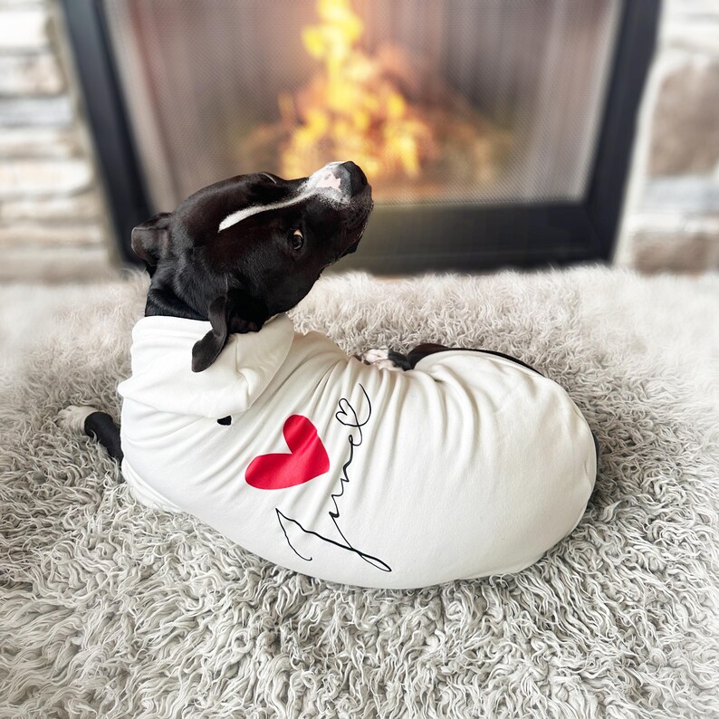 May include: A white dog hoodie featuring a red heart graphic and the cursive text "furever" printed in black. The garment has a hood and is worn by a black and white dog looking back over its shoulder toward a fireplace in the background.