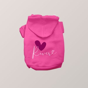 May include: A bright pink pet hoodie featuring a dark purple heart graphic above the white cursive text "River" which ends in a small heart outline. The garment includes a hood, short sleeves with ribbed cuffs, and a ribbed hem at the bottom edge.
