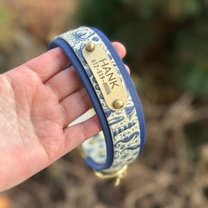 May include: A blue pet collar featuring a cream-colored fabric overlay with a nautical pattern of lighthouses, anchors, and sea creatures. A rectangular brass nameplate is attached, engraved with the name HANK and a phone number.