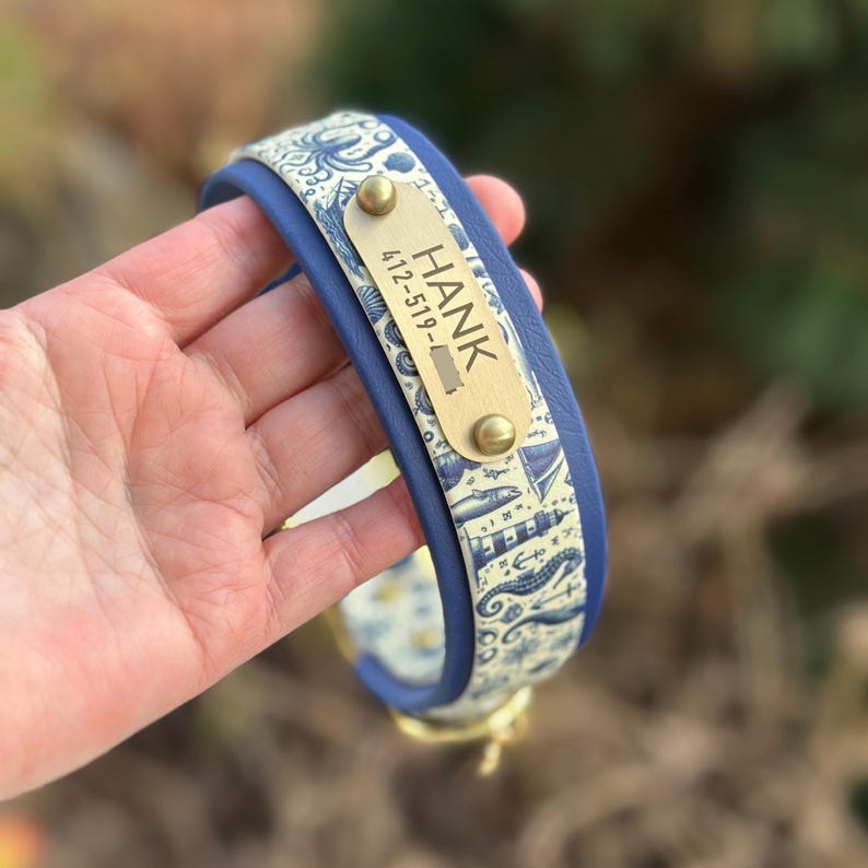 May include: A blue pet collar featuring a cream-colored fabric overlay with a nautical pattern of lighthouses, anchors, and sea creatures. A rectangular brass nameplate is attached, engraved with the name HANK and a phone number.