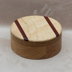 May include: Oval wooden box with a light-coloured lid featuring two diagonal dark brown stripes. The box's body is made of a darker wood, creating a contrast. This handcrafted box is suitable for storing small items.