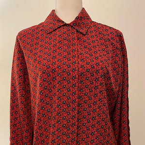 May include: A red button-down shirt with a small, repeating black and white floral pattern. The long-sleeved shirt has a classic collar and cuffs. The shirt is displayed on a mannequin.