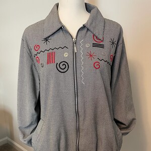 May include: A black and white houndstooth zip-up jacket featuring a pointed collar, long sleeves, and an elasticized hem. The front is decorated with embroidered red and black abstract shapes, including spirals, lines, and stars, along with small buttons.