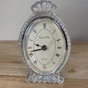 Puede incluir: Reloj de cuarzo transparente con esfera ovalada y números romanos. El reloj tiene un marco y una base decorativos transparentes y texturizados. La esfera es de color crema con las palabras "Crystal Clear" impresas.