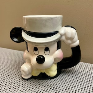 May include: A ceramic mug shaped like a cartoon mouse wearing a white top hat with a black band, a yellow bow tie, and black ears. The mug features a glossy finish with pink cheeks, a black nose, and a handle formed by a white gloved hand.