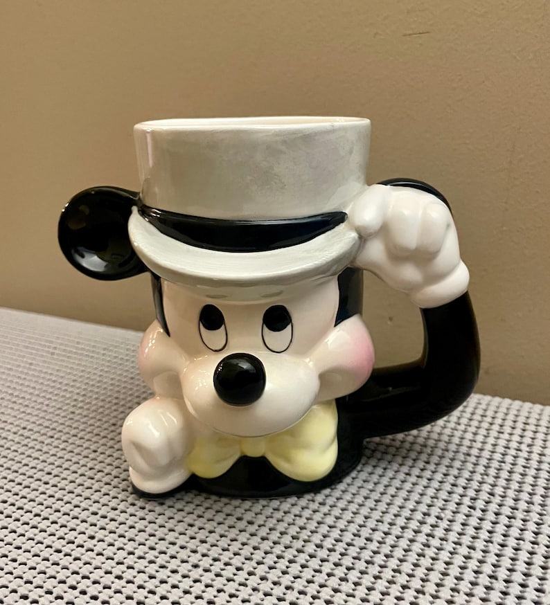 May include: A ceramic mug shaped like a cartoon mouse wearing a white top hat with a black band, a yellow bow tie, and black ears. The mug features a glossy finish with pink cheeks, a black nose, and a handle formed by a white gloved hand.
