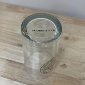 K&ouml;nnte beinhalten: Ein klares Trinkglas mit dem Text Guinness & Co., der auf den Boden gedruckt ist. An der Seite des Glases befindet sich ein teilweises kreisf&ouml;rmiges Logo mit dem Wort Guinness in schwarzer Schrift sowie weiterer Text am Rand.