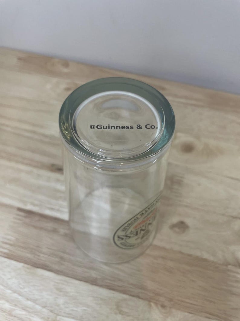 K&ouml;nnte beinhalten: Ein klares Trinkglas mit dem Text Guinness & Co., der auf den Boden gedruckt ist. An der Seite des Glases befindet sich ein teilweises kreisf&ouml;rmiges Logo mit dem Wort Guinness in schwarzer Schrift sowie weiterer Text am Rand.