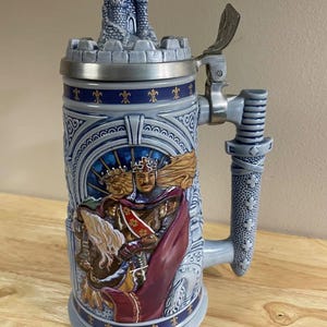 May include: A ceramic beer stein featuring a relief of a king in a red cape and crown. The lid is topped with a miniature castle, and the handle is shaped like a sword hilt. The body is light blue with dark blue fleur-de-lis accents and trim.