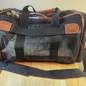 May include: A black pet carrier with brown faux leather trim and gold-tone hardware. The bag features a large mesh ventilation panel on the front, a top handle, and an adjustable black shoulder strap. The word SHERPA is printed on the zipper pull.