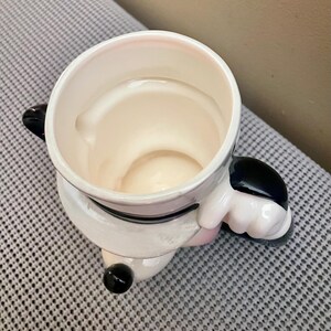 May include: A ceramic mug shaped like a cartoon character, featuring a white body with black accents on the ears and a small black nose. The mug has a wide, circular opening at the top and a sculpted handle on the side, showing a glossy finish.