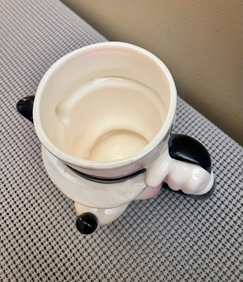May include: A ceramic mug shaped like a cartoon character, featuring a white body with black accents on the ears and a small black nose. The mug has a wide, circular opening at the top and a sculpted handle on the side, showing a glossy finish.