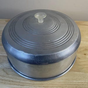 May include: A round, silver-colored cake carrier with a clear glass knob on the lid. The lid has concentric circular ridges. The carrier sits on a wooden surface.