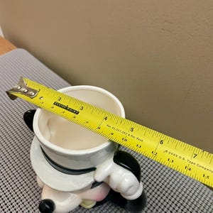 May include: A ceramic character mug featuring a white top hat with a black band. A yellow measuring tape is placed across the top opening, showing a diameter of approximately 4.5 inches. The mug has a white interior and black accents.