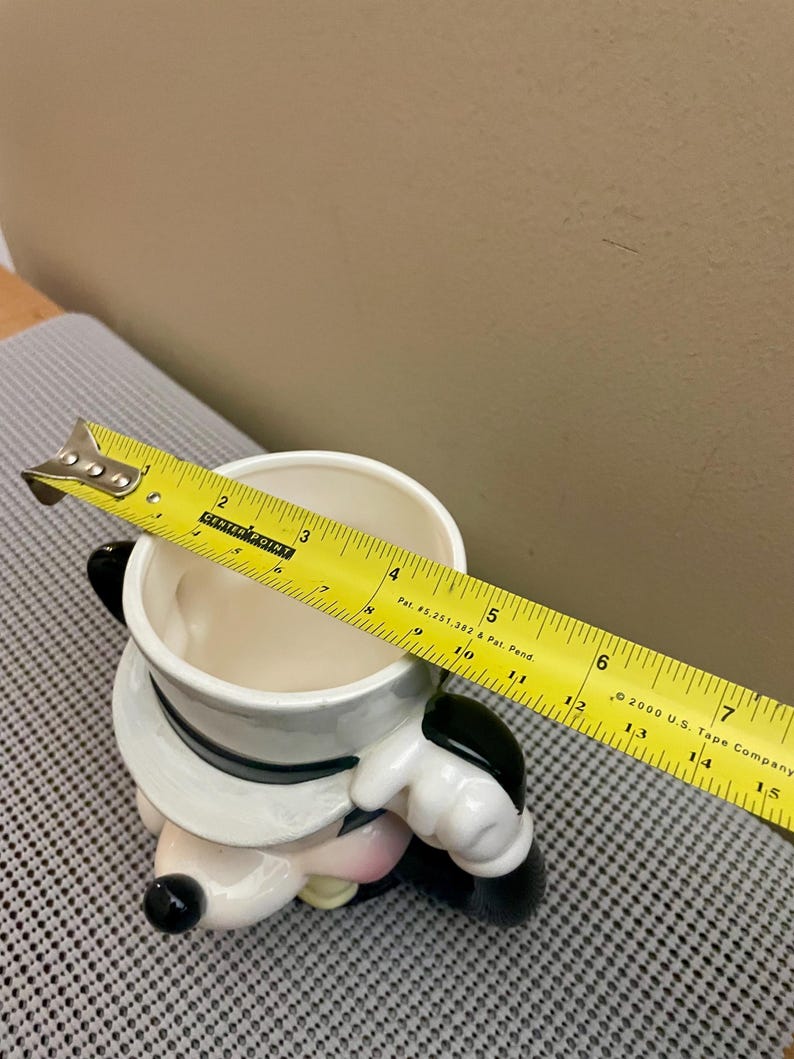 May include: A ceramic character mug featuring a white top hat with a black band. A yellow measuring tape is placed across the top opening, showing a diameter of approximately 4.5 inches. The mug has a white interior and black accents.