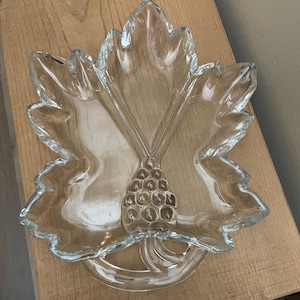 Puede incluir: Fuente de vidrio transparente en forma de hoja con borde texturizado y diseño de racimo de uvas en el centro. La fuente está diseñada para parecerse a una hoja grande, con vetas detalladas y un borde festoneado. Una pieza decorativa.