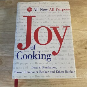 May include: A cookbook titled "Joy of Cooking" with a red bookmark. The cover features the title in large red letters, with the words "All New All Purpose" above. The book lists various recipes and authors.