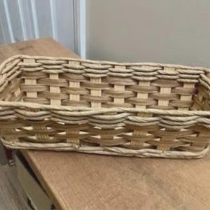 May include: A rectangular woven basket with a natural, light brown color. The basket is made of interwoven strips of material, creating a textured surface. It sits on a wooden surface.