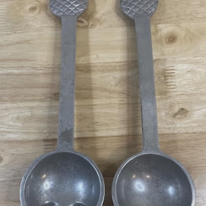 Könnte beinhalten: Zwei silberfarbene Servierutensilien. Eines ist ein Löffel, das andere eine Gabel. Beide haben ein Ananas-Design am oberen Ende des Griffs. Löffel und Gabel sind aus Metall und liegen auf einer Holzoberfläche.