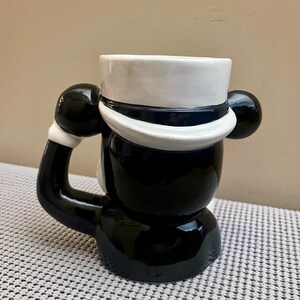 May include: A ceramic mug shaped like a character wearing a white top hat with a dark blue band. The mug features a glossy black finish on the body and a handle designed as a black arm with a white cuff, extending from the side of the figure.