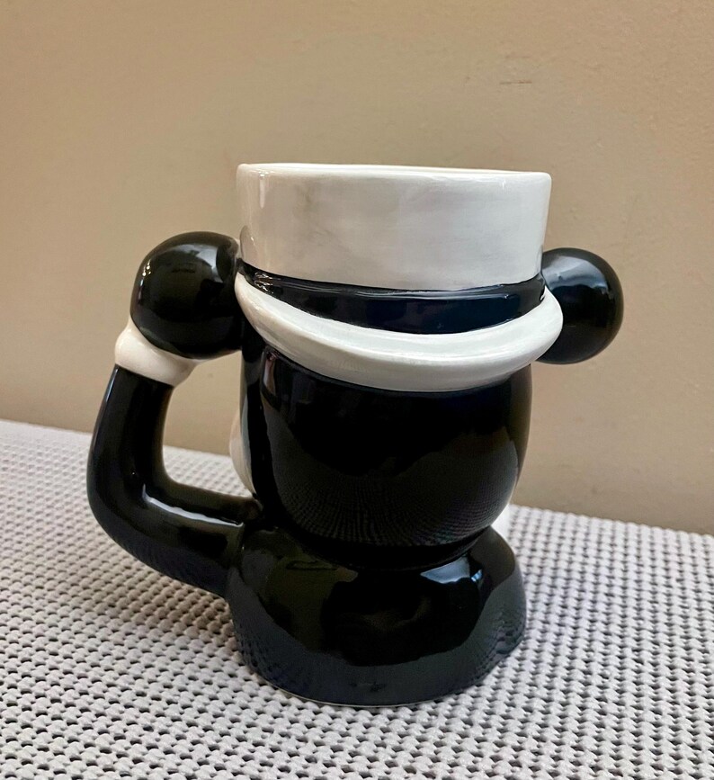 May include: A ceramic mug shaped like a character wearing a white top hat with a dark blue band. The mug features a glossy black finish on the body and a handle designed as a black arm with a white cuff, extending from the side of the figure.