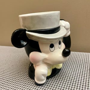 May include: A ceramic mug shaped like a cartoon mouse head wearing a white top hat with a black band. The mouse has black ears, a white face with a black nose, pink cheeks, and a yellow bowtie. The mug features a glossy finish and a handle.