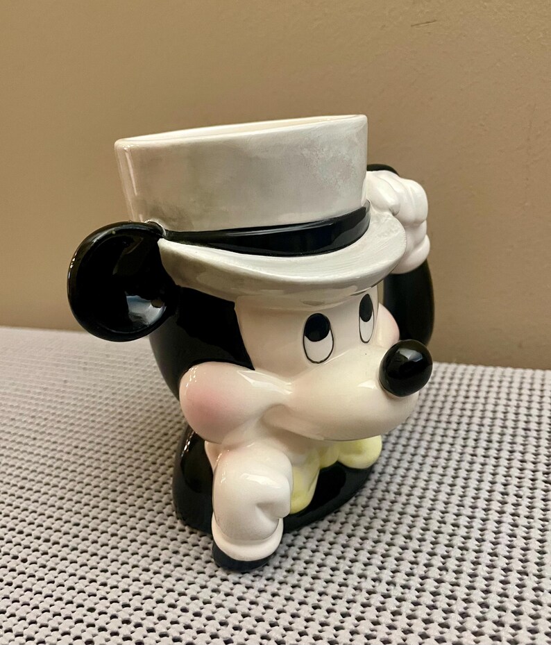 May include: A ceramic mug shaped like a cartoon mouse head wearing a white top hat with a black band. The mouse has black ears, a white face with a black nose, pink cheeks, and a yellow bowtie. The mug features a glossy finish and a handle.