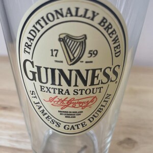 K&ouml;nnte beinhalten: Ein klares Bierglas mit einem runden, cremefarbenen Etikett, das das schwarze Guinness-Logo, eine Harfe, den Text TRADITIONALLY BREWED, 1759, EXTRA STOUT, die Unterschrift Arth Guinness sowie ST JAMES'S GATE DUBLIN zeigt.