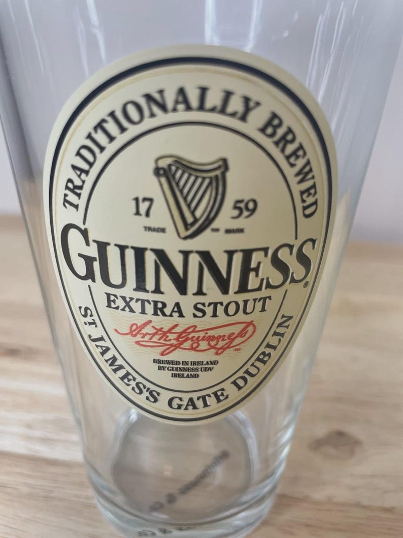K&ouml;nnte beinhalten: Ein klares Bierglas mit einem runden, cremefarbenen Etikett, das das schwarze Guinness-Logo, eine Harfe, den Text TRADITIONALLY BREWED, 1759, EXTRA STOUT, die Unterschrift Arth Guinness sowie ST JAMES'S GATE DUBLIN zeigt.