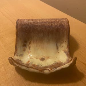 May include: A ceramic soap dish with a curved back and a shallow, rounded base. The dish has a mottled glaze in shades of brown, cream, and tan. The soap dish is designed to hold a bar of soap.