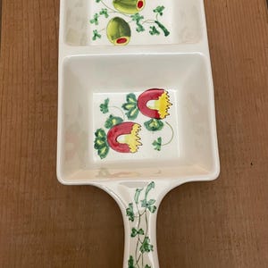 May include: A white ceramic serving dish with two square compartments and a handle. The dish is decorated with green olives and red and yellow fruit designs. The handle features green leaf patterns.