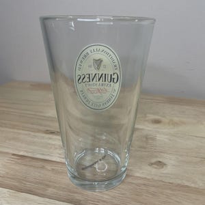 K&ouml;nnte beinhalten: Ein klares Glas-Pintglas mit einer konischen Form und einem dicken Boden. Auf der Vorderseite befindet sich ein wei&szlig;es, ovales Logo mit dem Schriftzug Guinness Extra Stout und einem Harfen-Emblem, das deutlich auf dem Glas zu sehen ist.