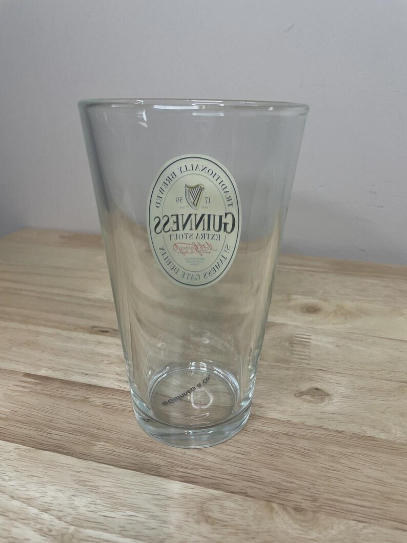 K&ouml;nnte beinhalten: Ein klares Glas-Pintglas mit einer konischen Form und einem dicken Boden. Auf der Vorderseite befindet sich ein wei&szlig;es, ovales Logo mit dem Schriftzug Guinness Extra Stout und einem Harfen-Emblem, das deutlich auf dem Glas zu sehen ist.