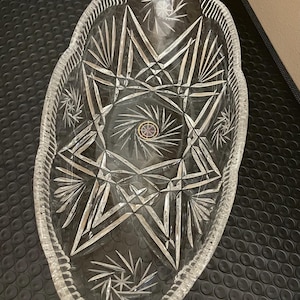 May include: An oval clear glass serving dish featuring intricate cut patterns including starburst motifs and geometric lines. The center of the dish displays a small circular sticker with a gold border and a multi-colored star design.