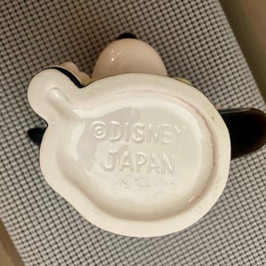 May include: The underside of a ceramic mug featuring the embossed text Disney and Japan. The base is off-white with a smooth, glossy finish, showing a portion of the dark handle and the rounded shape of the mug extending from the bottom edge.