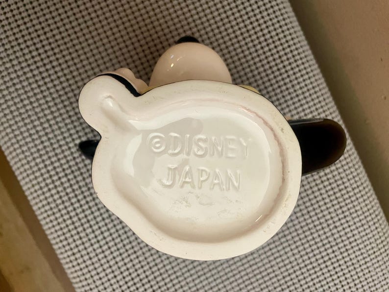 May include: The underside of a ceramic mug featuring the embossed text Disney and Japan. The base is off-white with a smooth, glossy finish, showing a portion of the dark handle and the rounded shape of the mug extending from the bottom edge.