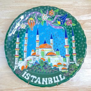 Può includere: Piatto decorativo in ceramica con una dettagliata illustrazione di una moschea a Istanbul. Il piatto ha un bordo verde con motivi floreali e la scritta "ISTANBUL" in bianco. La moschea è raffigurata con pareti bianche, cupole arancioni e minareti blu.