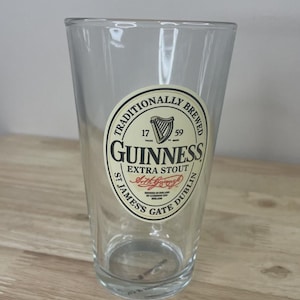 K&ouml;nnte beinhalten: Ein klares Glas-Pint mit einem kreisf&ouml;rmigen, cremefarbenen Etikett, das den schwarzen Text TRADITIONALLY BREWED GUINNESS EXTRA STOUT ST JAMES'S GATE DUBLIN, ein Harfensymbol, die Jahreszahlen 1759 und eine rote Signaturgrafik auf der Vorderseite zeigt.