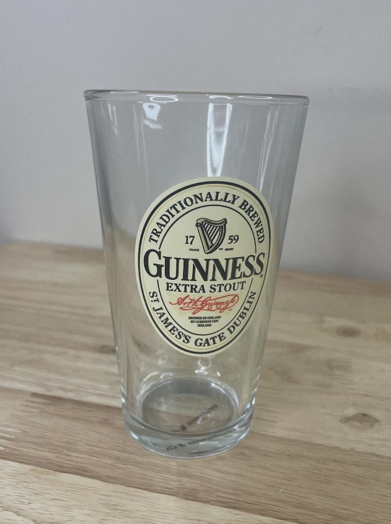 K&ouml;nnte beinhalten: Ein klares Glas-Pint mit einem kreisf&ouml;rmigen, cremefarbenen Etikett, das den schwarzen Text TRADITIONALLY BREWED GUINNESS EXTRA STOUT ST JAMES'S GATE DUBLIN, ein Harfensymbol, die Jahreszahlen 1759 und eine rote Signaturgrafik auf der Vorderseite zeigt.
