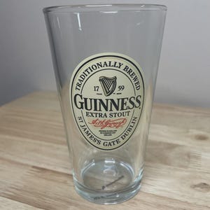 K&ouml;nnte beinhalten: Ein klares Glas-Pintglas mit einem kreisf&ouml;rmigen cremefarbenen Etikett mit dem Text TRADITIONALLY BREWED GUINNESS EXTRA STOUT ST JAMES'S GATE DUBLIN, einem Harfensymbol, den Jahren 1759 und der Unterschrift von Arthur Guinness in roter Tinte.