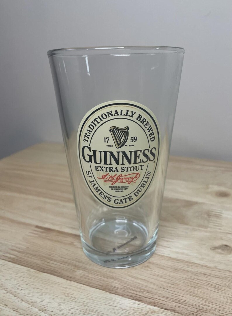 K&ouml;nnte beinhalten: Ein klares Glas-Pintglas mit einem kreisf&ouml;rmigen cremefarbenen Etikett mit dem Text TRADITIONALLY BREWED GUINNESS EXTRA STOUT ST JAMES'S GATE DUBLIN, einem Harfensymbol, den Jahren 1759 und der Unterschrift von Arthur Guinness in roter Tinte.