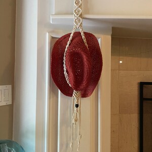 May include: A terracotta colored woven hat hanging from a white macrame hat holder with a black bead accent.