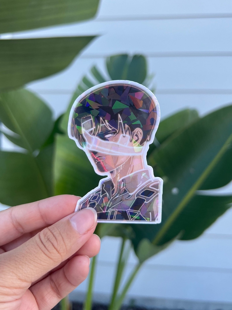 Peut inclure: Un autocollant holographique repr&eacute;sentant un personnage de l'anime L'Attaque des Titans. Le personnage a les cheveux noirs et porte un uniforme militaire. L'autocollant est iridescent et a une finition scintillante.
