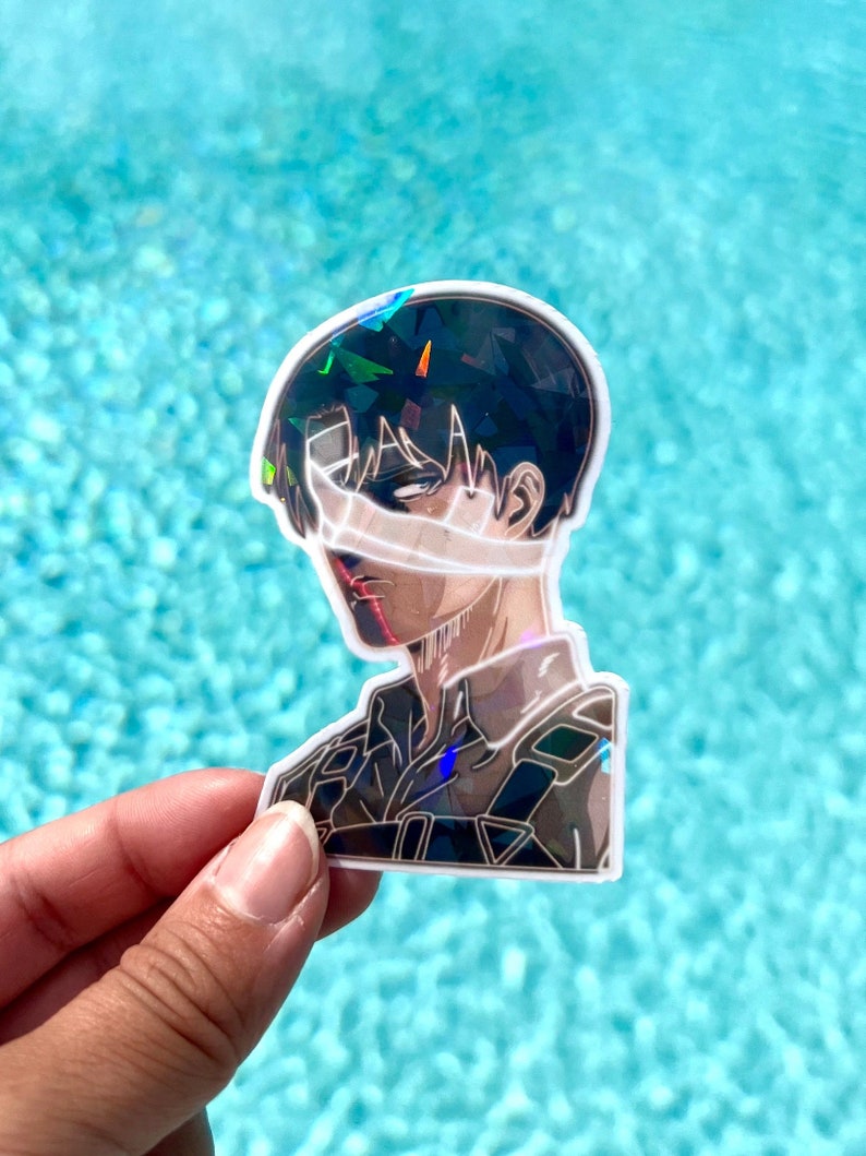 Peut inclure: Un autocollant holographique repr&eacute;sentant un personnage de l'anime L'Attaque des Titans. Le personnage a les cheveux noirs et porte un uniforme militaire. L'autocollant est sur un fond bleu.