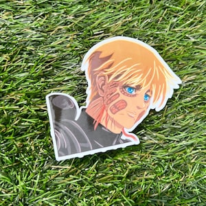May include: A sticker featuring a cartoon character with blond hair and blue eyes. The character has a distressed look on their face and is wearing a black and gray outfit. The sticker is on a green background.