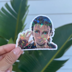 Puede incluir: Una pegatina hologr&aacute;fica con un personaje del anime Ataque a los Titanes. El personaje tiene el pelo oscuro y los ojos azules y lleva una chaqueta oscura. La pegatina est&aacute; hecha de un material de vinilo transparente y tiene un acabado hologr&aacute;fico.