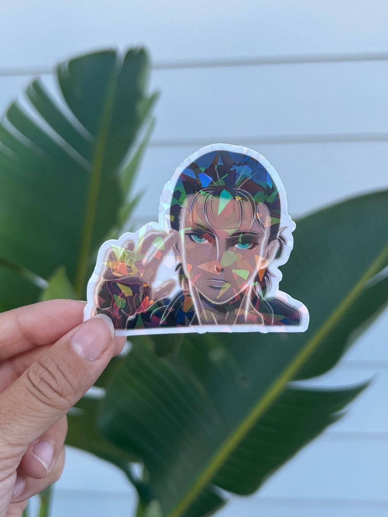 Puede incluir: Una pegatina hologr&aacute;fica con un personaje del anime Ataque a los Titanes. El personaje tiene el pelo oscuro y los ojos azules y lleva una chaqueta oscura. La pegatina est&aacute; hecha de un material de vinilo transparente y tiene un acabado hologr&aacute;fico.