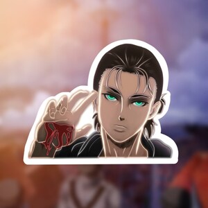 Puede incluir: Una pegatina con un personaje del anime Ataque a los Titanes. El personaje tiene el pelo oscuro, los ojos verdes y una mano ensangrentada levantada frente a su cara.